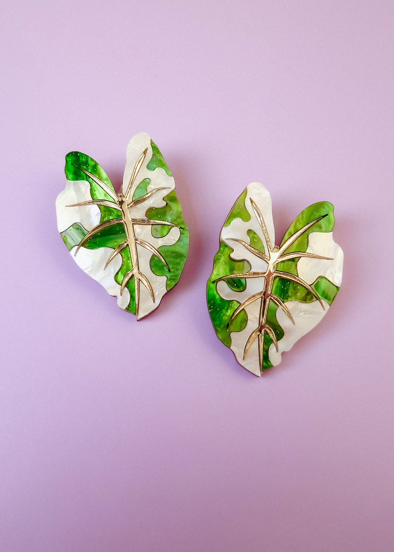 Two decorative pins shaped like monstera leaves, inspired by hand-crafted jewelry, feature a mix of green and white colors with gold vein detailing, and are placed on a light purple background.