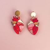 A pair of gold and pink Anatomical Heart Earrings, crafted in acrylic with detailed vein patterns and a unique style, displayed on a light pink background.