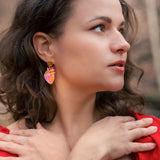 A woman with wavy brown hair and a tattooed forearm crosses her hands over her chest. She wears a red dress, bold style pink statement earrings, and stands outdoors among brown autumn leaves, looking to the side.