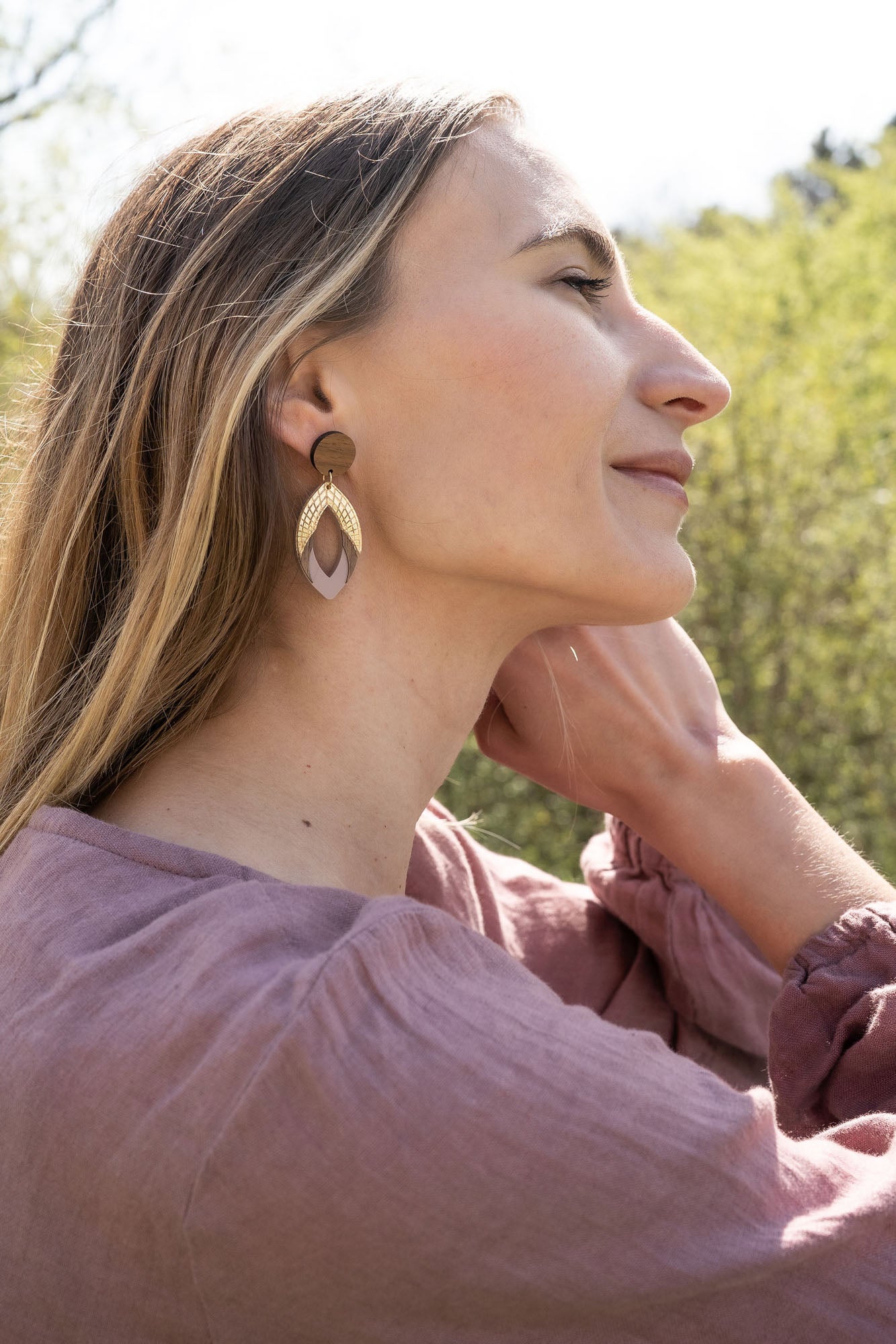 A woman with long hair wearing a mauve top and large handcrafted leaf-shaped earrings sits outdoors, tilting her head back and touching her neck, with greenery and trees blurred in the background.