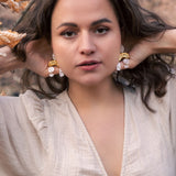 A woman with wavy brown hair poses outdoors among dry autumn leaves, wearing a light beige dress and boho chic dangle earrings adorned with rhinestones, her hands touching her hair and an intense, confident expression on her face.