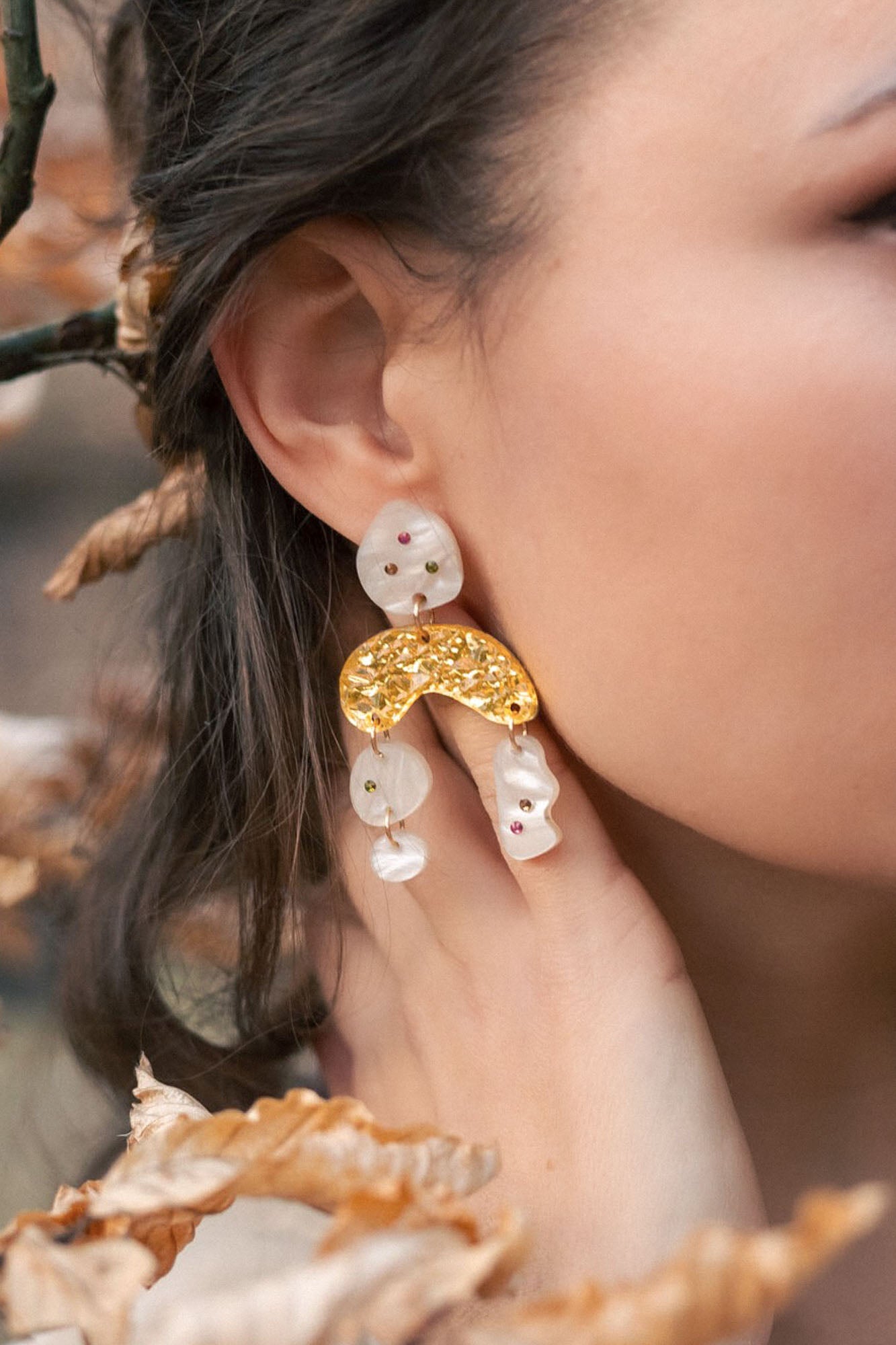 A close-up of a womans ear wearing unique boho chic dangle earrings with gold, pearl-like abstract shapes, and subtle rhinestones. Her hand touches her neck, surrounded by brown autumn leaves and branches.