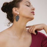 A woman with closed eyes and an elegant pose wears a burgundy off-the-shoulder top, a delicate gold necklace, and Calathea Ornata Earrings—handcrafted earrings inspired by tropical nature jewelry. Her dark hair is tied up as she holds a straw hat on her lap.