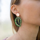 Close-up of a woman wearing Calathea Medallion Earrings—handcrafted statement jewelry in green and black with a gold edge. She has light brown hair, a mauve top, and only the right side of her face is visible.