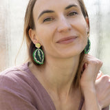A woman with light brown hair, wearing a mauve blouse and green Calathea Medallion Earrings, sits by a sunlit window, smiling gently at the camera, her nature-inspired jewelry adding a unique touch to her look.