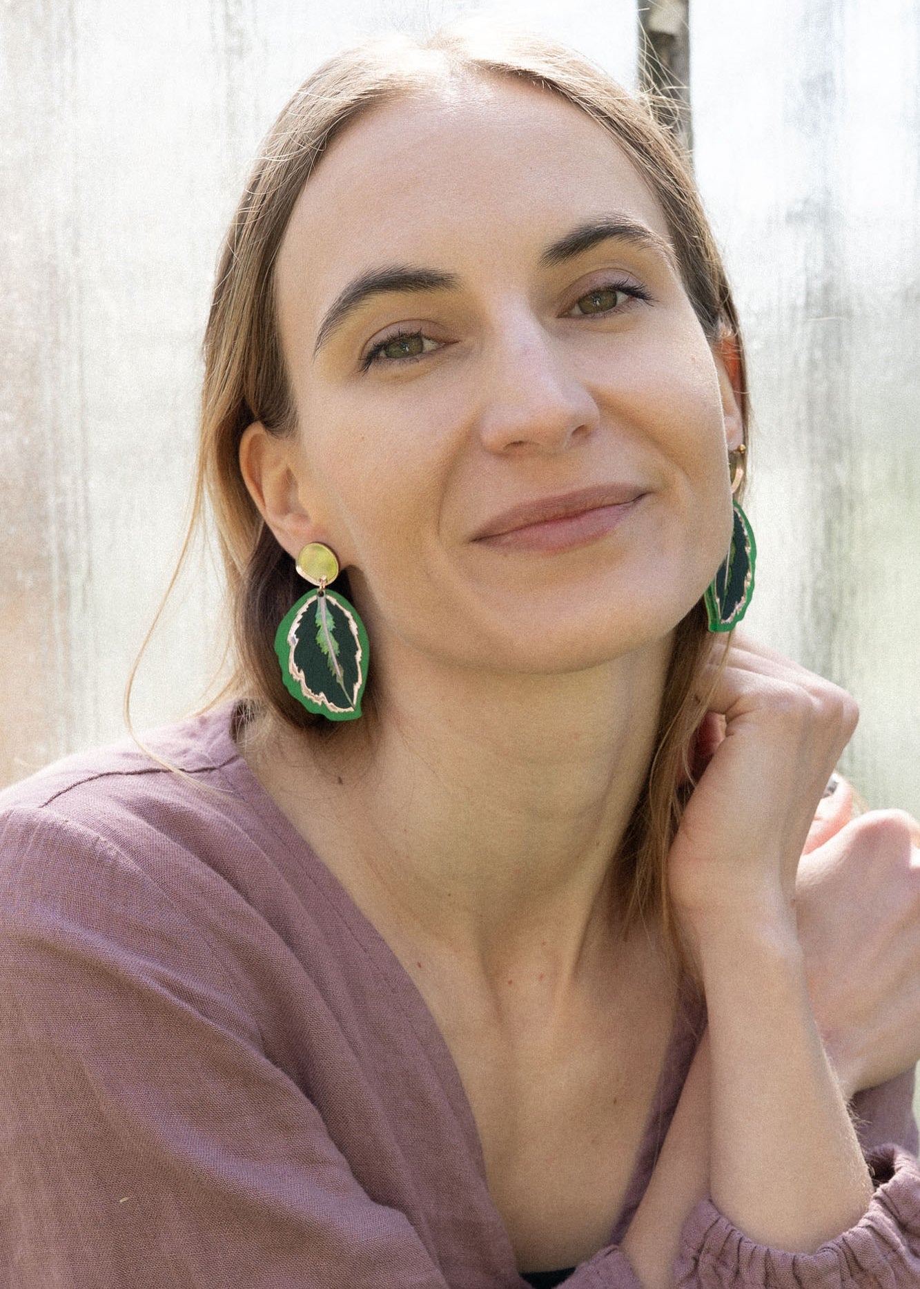 A woman with light brown hair, wearing a mauve blouse and green Calathea Medallion Earrings, sits by a sunlit window, smiling gently at the camera, her nature-inspired jewelry adding a unique touch to her look.
