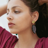 A woman with wavy brown hair pulled back wears a burgundy top, a delicate gold necklace, and Calathea Ornata Earrings—handcrafted earrings inspired by tropical nature jewelry—featuring green beads and a black-and-white leaf design.