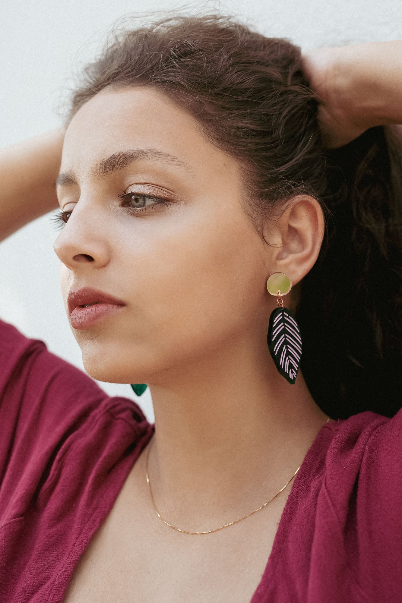A woman with wavy brown hair pulled back wears a burgundy top, a delicate gold necklace, and Calathea Ornata Earrings—handcrafted earrings inspired by tropical nature jewelry—featuring green beads and a black-and-white leaf design.