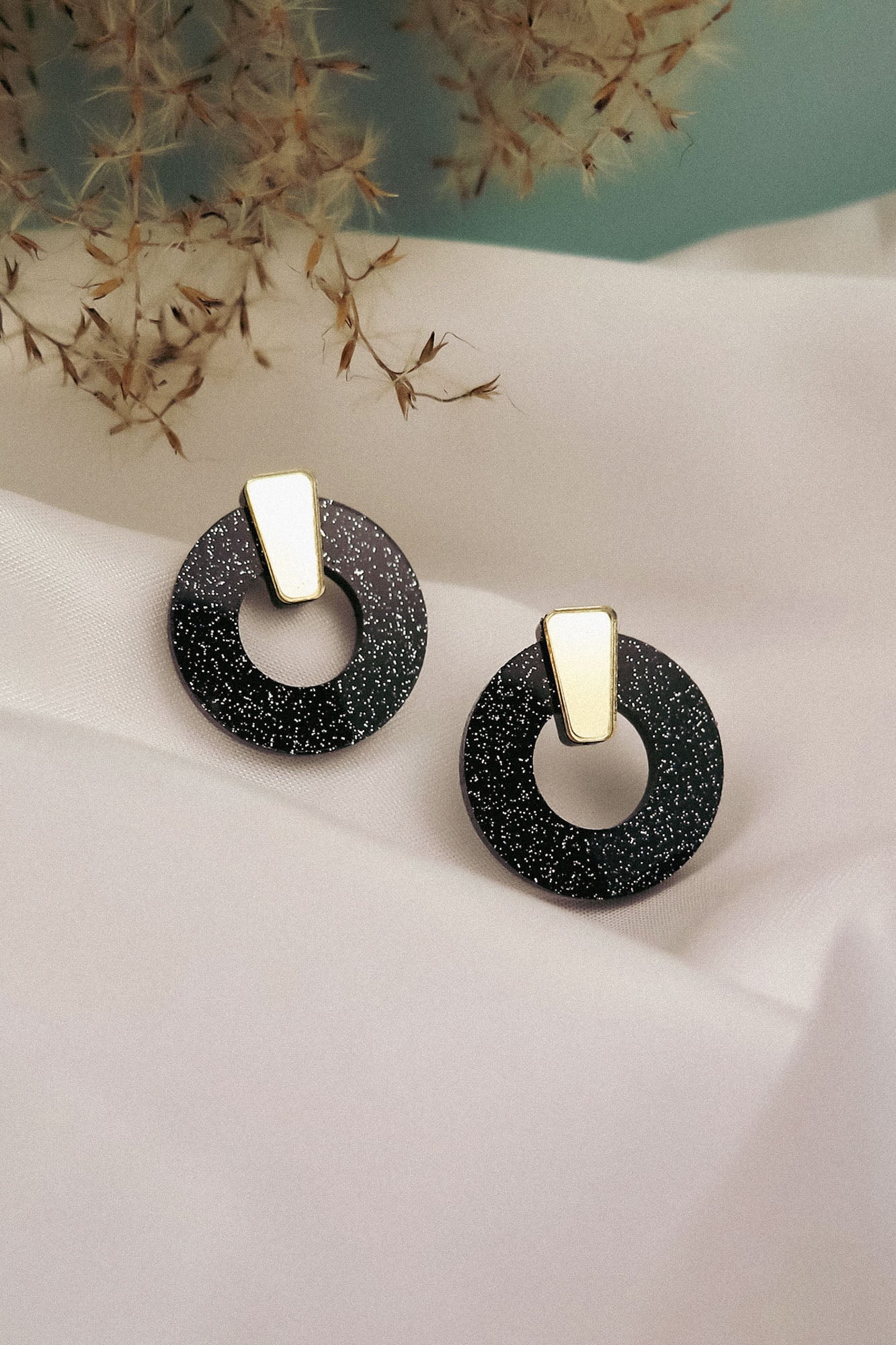 A pair of minimalist circle stud earrings in black acrylic glass with white speckles and gold triangular studs, displayed on white fabric with dried grasses in the background. Hypoallergenic for comfortable, everyday wear.