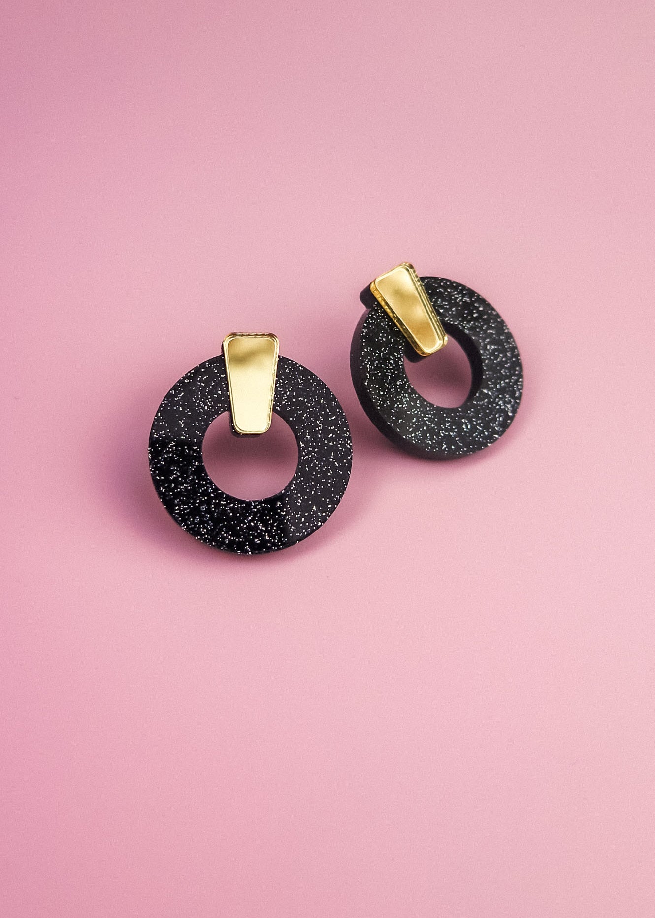 A pair of minimalist circle stud earrings, crafted from hypoallergenic acrylic glass with white speckles and gold rectangular accents, are displayed on a pink background.