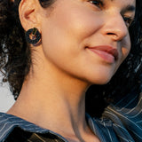 A woman with curly hair smiles gently, wearing a striped shirt and large round hypoallergenic earrings, with one hand behind her head, in soft natural light.