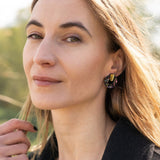 A woman with long, light brown hair stands outdoors, wearing a dark coat, mauve top, and minimalist circle stud earrings. She lightly touches her hair and looks calmly at the camera against green foliage and a bright, sunlit sky.