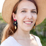 A woman with light brown hair wearing a wide-brimmed straw hat, artisan dangle earrings, and a white top smiles outdoors, surrounded by greenery and sunlight.
