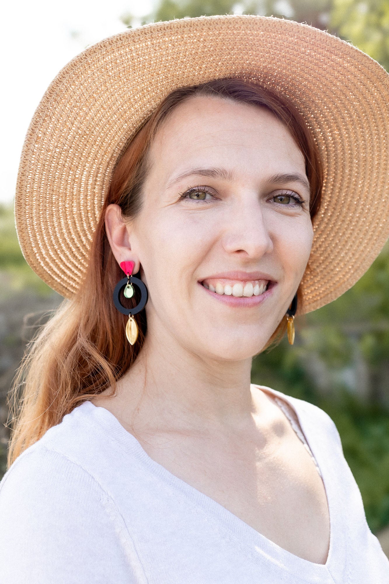 A woman with light brown hair wearing a wide-brimmed straw hat, artisan dangle earrings, and a white top smiles outdoors, surrounded by greenery and sunlight.