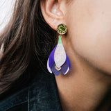 Close-up of a person wearing Crocus Flower Earrings—a handcrafted, statement accessory shaped like a flower petal in purple, white, and gold. The person has long brown hair and is dressed in a dark-colored top.