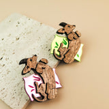 Two playful brooches shaped like reclining dachshunds, each resting on a colorful pillow—one pink and one neon green—placed on a textured stone surface. A charming touch for any dog accessory collection or fans of unique brooches.