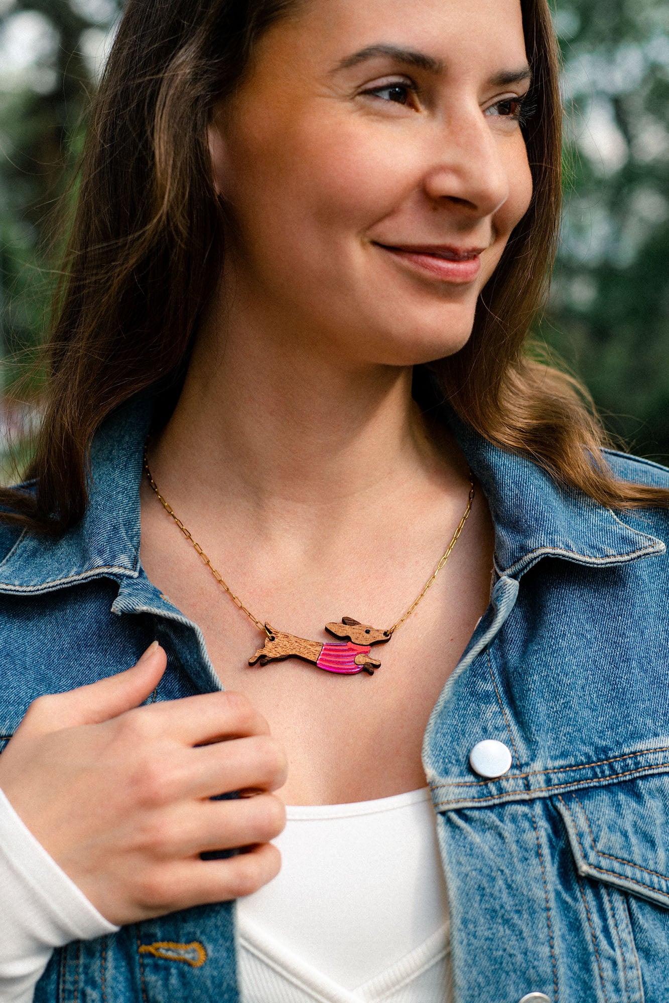 A woman wearing a denim jacket and white top smiles outdoors. Her long brown hair frames a colorful, handcrafted necklace featuring a Dachshund dog design made from sustainable wood.