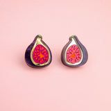 Two enamel pins shaped like halved figs with bright pink and red centers rest on a light pink background, perfect for fans of fig jewelry or those who love unique laser-cut studs.