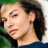 A woman with curly dark hair wears a blue top and bold hand-painted earrings in red, gold, and black. She is looking slightly to the side, partially surrounded by green leaves, with soft natural light on her face.