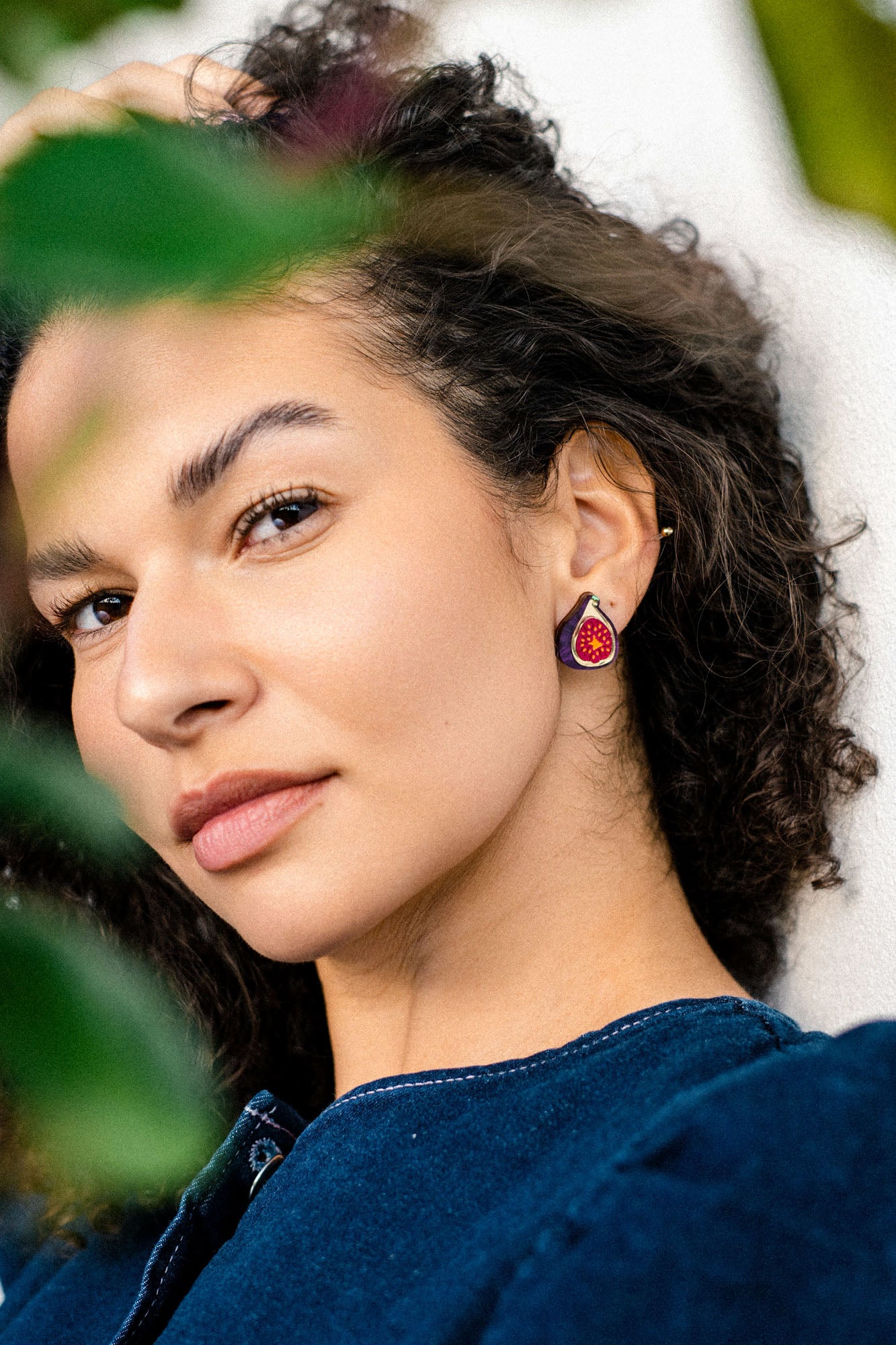 A woman with curly dark hair wears a blue top and bold hand-painted earrings in red, gold, and black. She is looking slightly to the side, partially surrounded by green leaves, with soft natural light on her face.