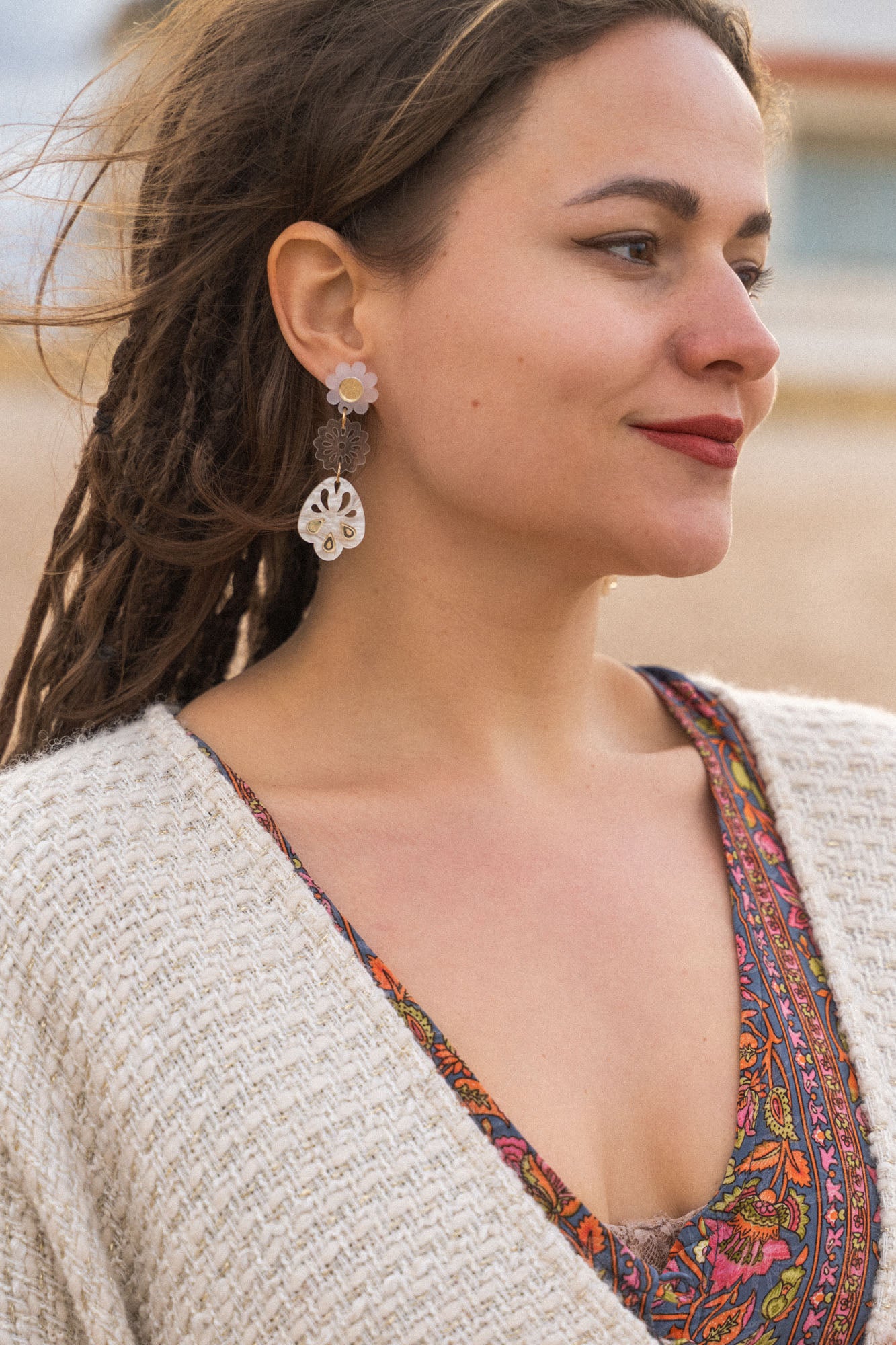 A woman with long braided hair stands outdoors, wearing a patterned dress and a cream knit sweater. She has handcrafted boho earrings and is smiling softly, looking to the side. The background appears to be a beach or sandy area.