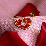A handcrafted gold Fragment of Love Necklace with a geometric red heart pendant rests on soft pink fabric, surrounded by scattered red rose petals.