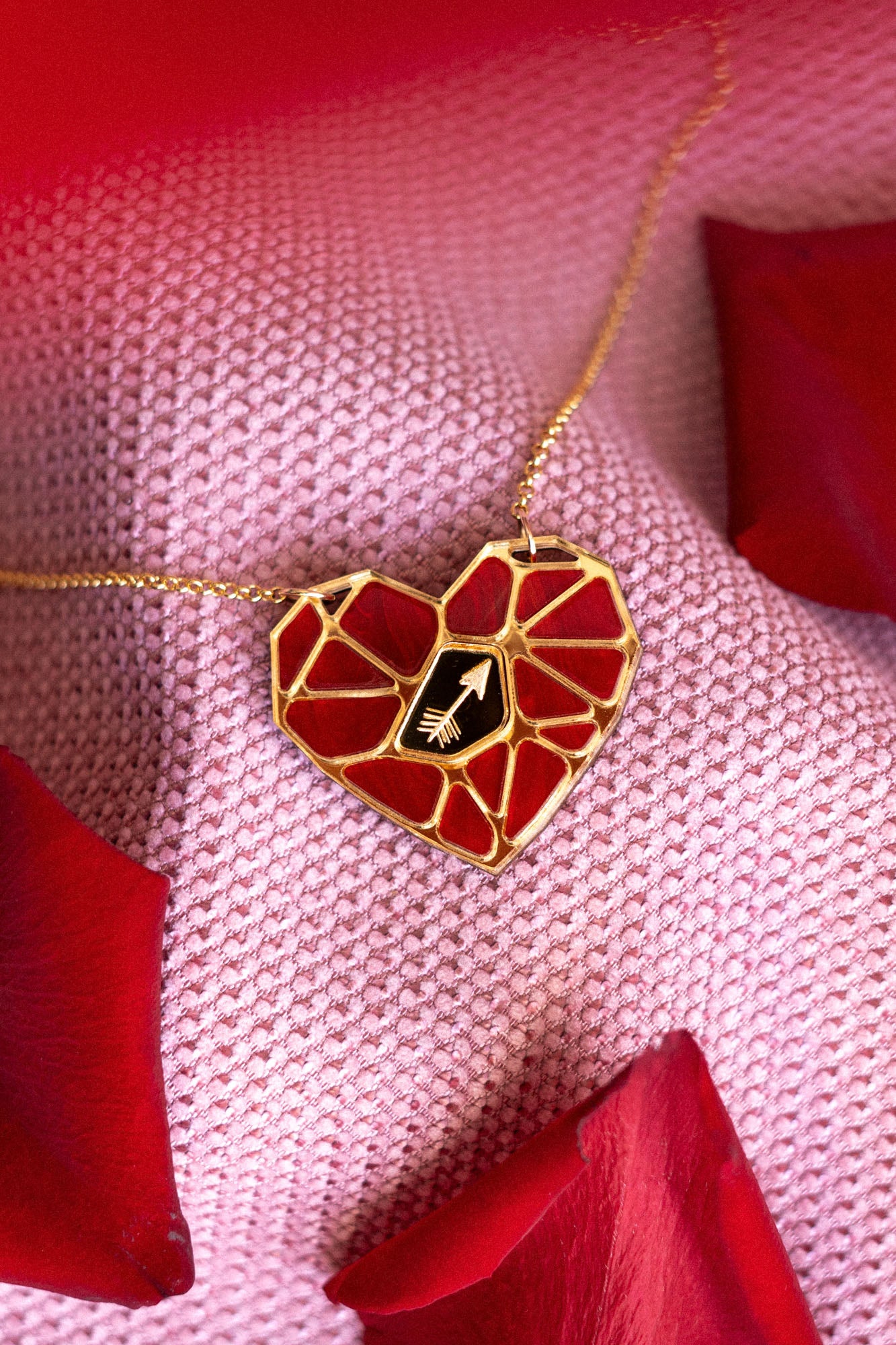 A handcrafted gold heart pendant, featuring a red mosaic design and a small white hand symbol, rests on pink textured fabric amid red rose petals—a striking Fragment of Love Necklace.