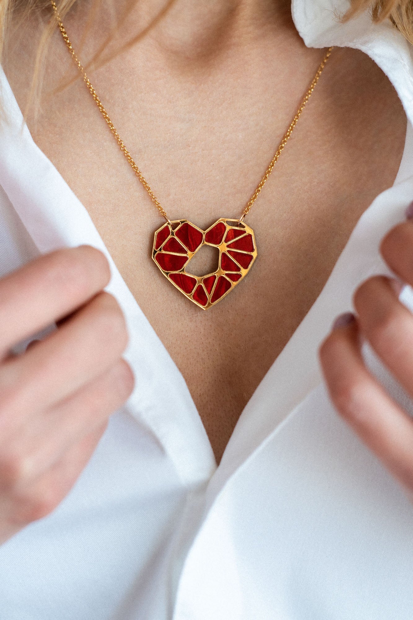 A close-up of a person in a white shirt, holding the collar open to reveal a handcrafted gold necklace with a geometric red heart pendant—an elegant Fragment of Love Necklace displayed around their neck.