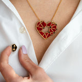 A person wearing a white shirt displays the handcrafted Fragment of Love Necklace featuring a red, geometric heart pendant and holds a small black pin with a gold arrow design. Only the lower face and hand are visible.