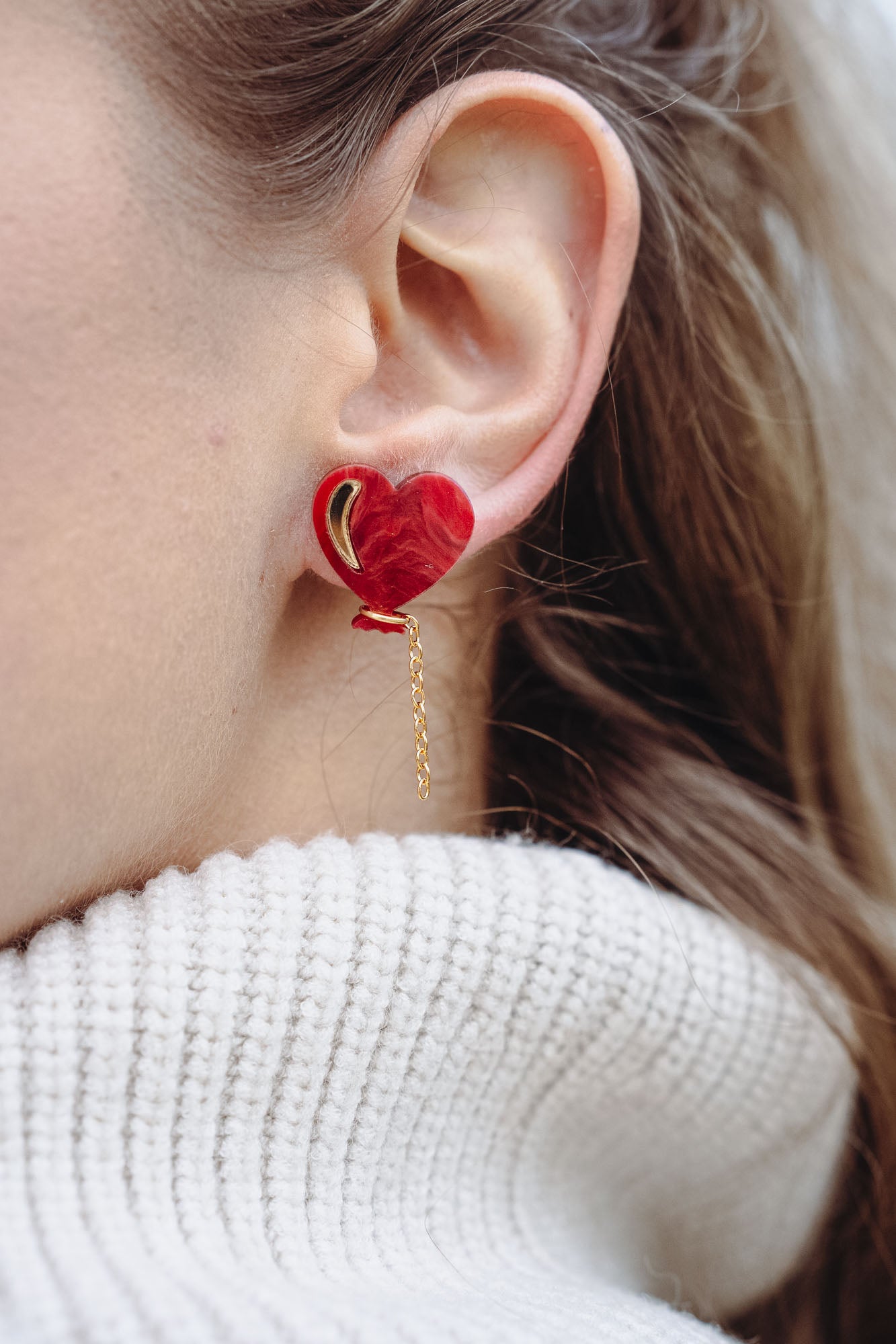Close-up of a woman's ear wearing playful Heart Balloon Studs—red heart-shaped earrings with a small gold chain. Part of her face and blonde hair is visible, complemented by a white ribbed turtleneck sweater.