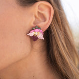 A close-up of a person’s ear wearing Hellebore Stud Earrings, handcrafted floral jewelry with pink petals and a purple center. The person has long light brown hair and is outdoors in natural light, showcasing these unique accessories.