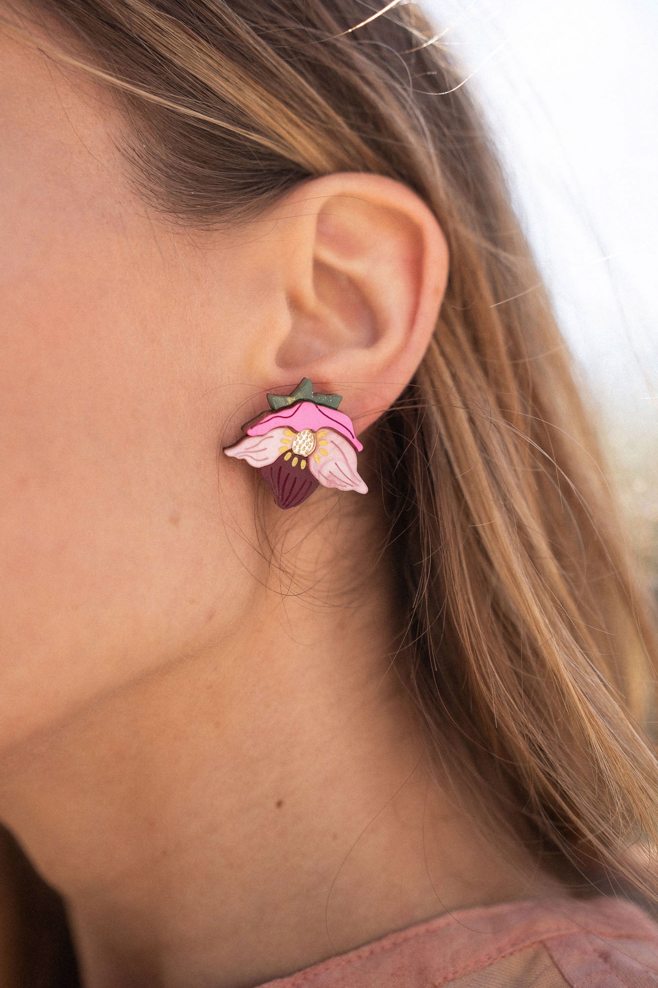 A close-up of a person’s ear wearing Hellebore Stud Earrings, handcrafted floral jewelry with pink petals and a purple center. The person has long light brown hair and is outdoors in natural light, showcasing these unique accessories.