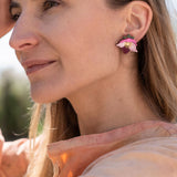 A woman with long hair wearing a peach-colored blouse and handcrafted floral jewelry stands outdoors, gently touching a tree branch and smiling softly in natural sunlight.