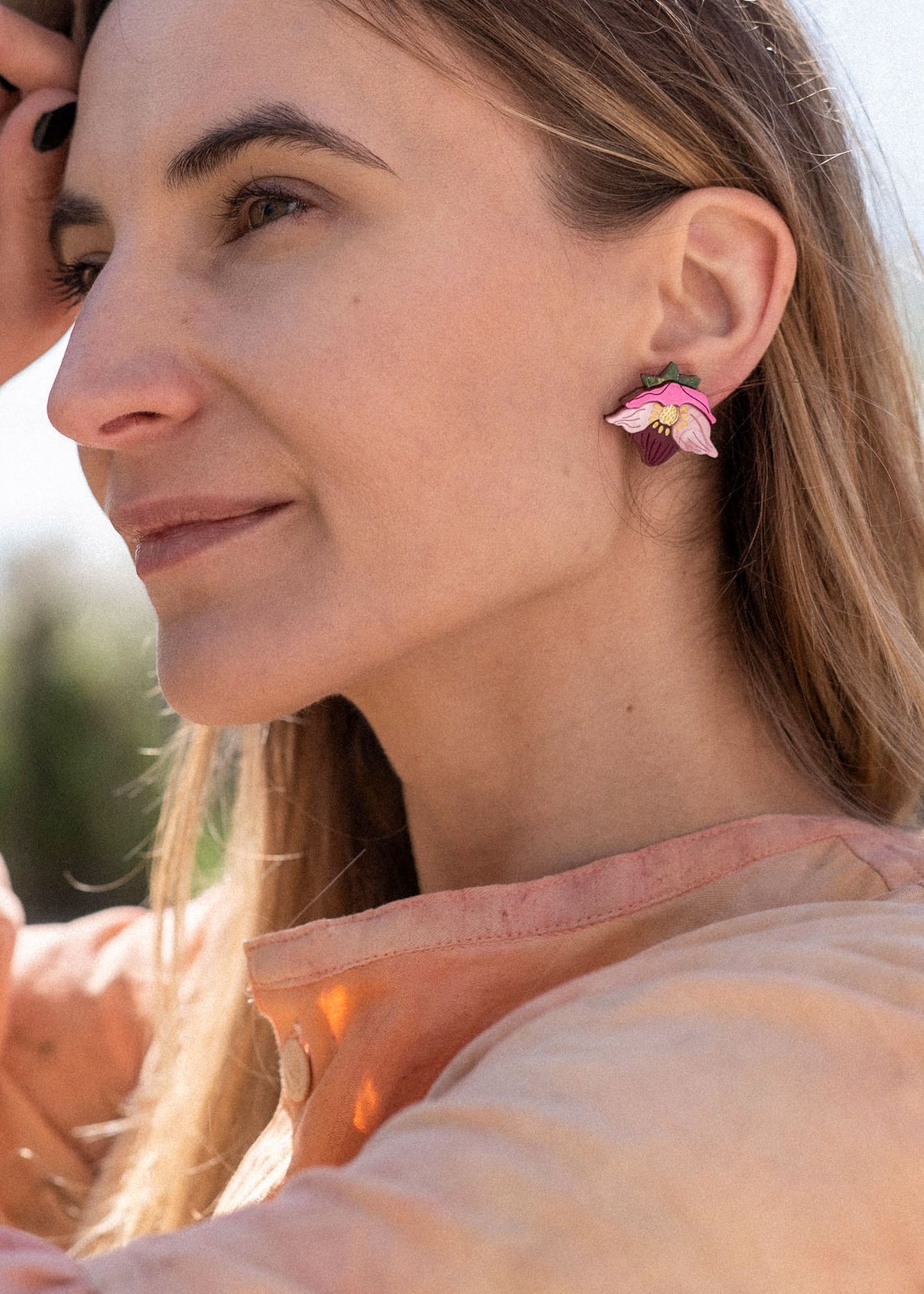 A woman with long hair wearing a peach-colored blouse and handcrafted floral jewelry stands outdoors, gently touching a tree branch and smiling softly in natural sunlight.