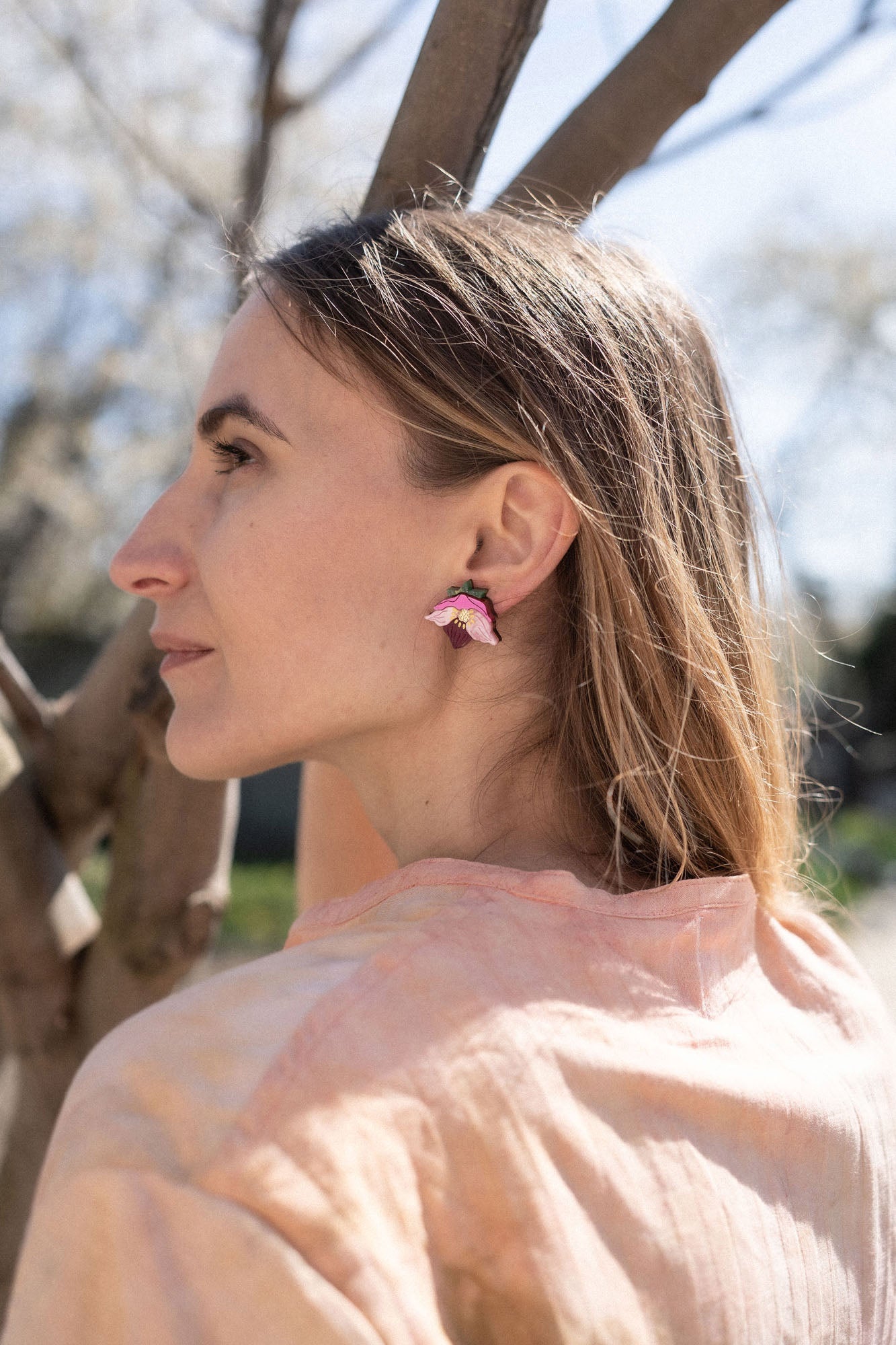 A woman with light brown hair stands outdoors near tree branches, wearing a light pink shirt and unique accessories—colorful, handcrafted floral jewelry that includes striking pink Hellebore Stud Earrings—as she gazes thoughtfully into the distance on a sunny day.