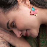 A woman with long brown hair lies on her side outdoors, displaying vibrant Hibiscus Earrings and a botanical tattoo on her arm. She is wearing a green patterned garment and appears calm and relaxed, making a tropical statement.
