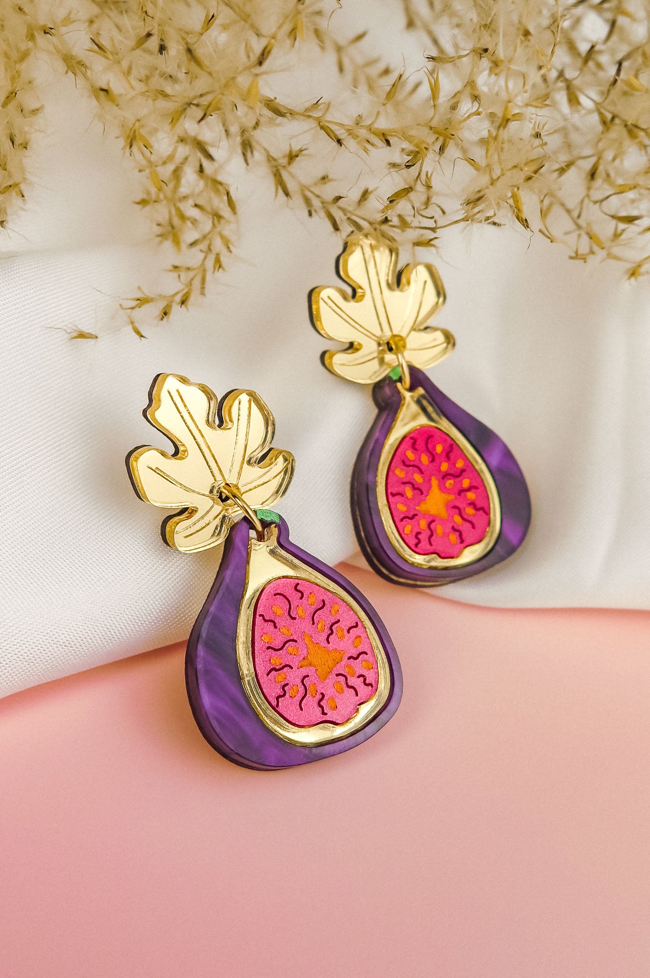 A pair of handcrafted Fig Earrings with gold mirror leaves, purple edges, and a pink center, displayed on a soft white and pink background with dried flowers.