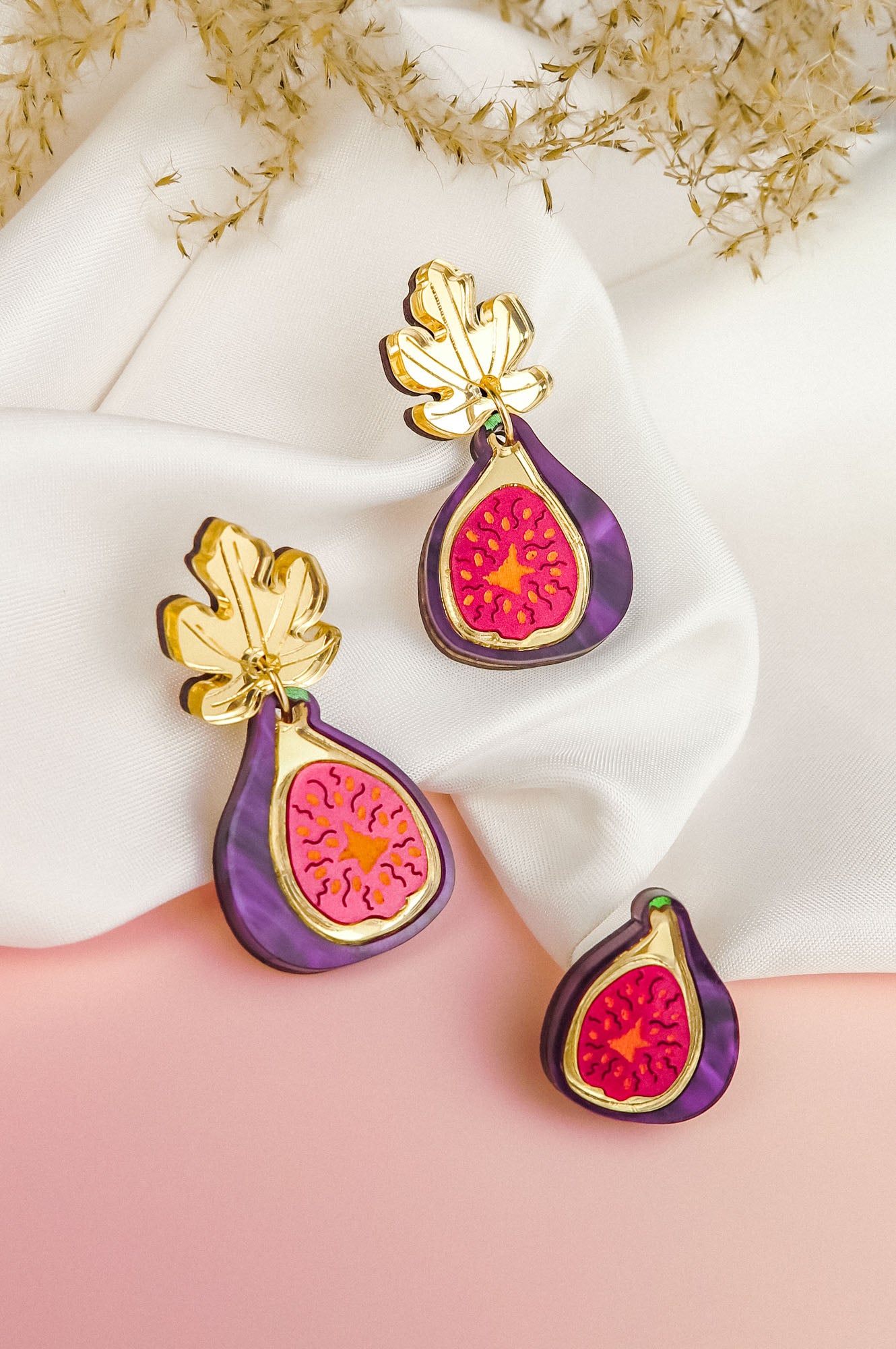 Three handcrafted Fig Earrings, featuring gold mirror leaves and vibrant purple and pink detailing, are displayed on white fabric and a pink surface, with dried foliage in the background.