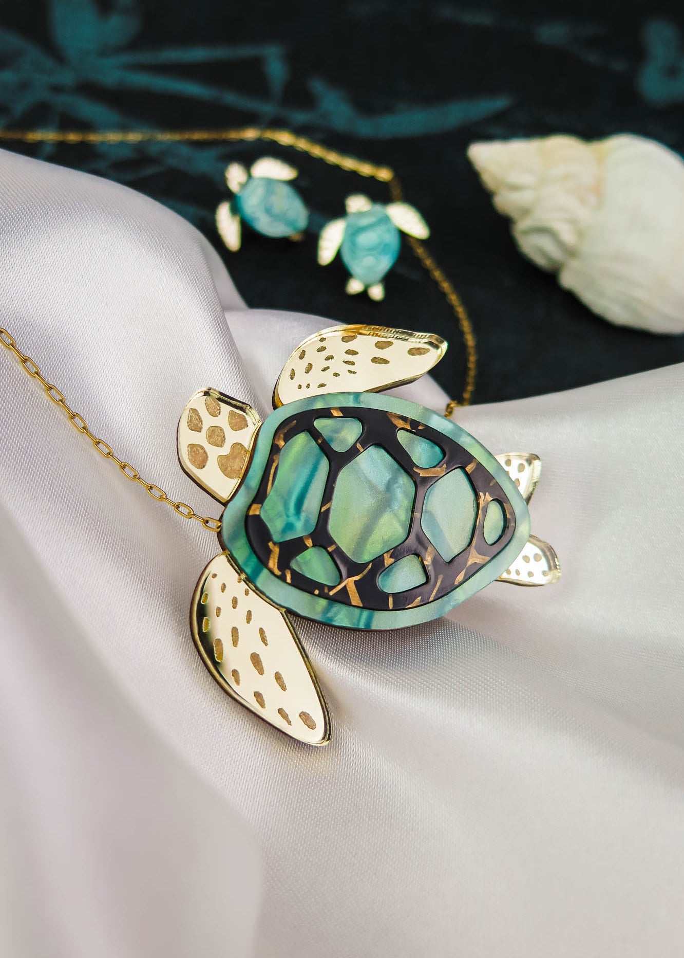 A gold and green Sea Turtle Necklace pendant, handcrafted jewelry with intricate details, is displayed on silky white fabric alongside a matching smaller turtle necklace in the background and a seashell nearby.