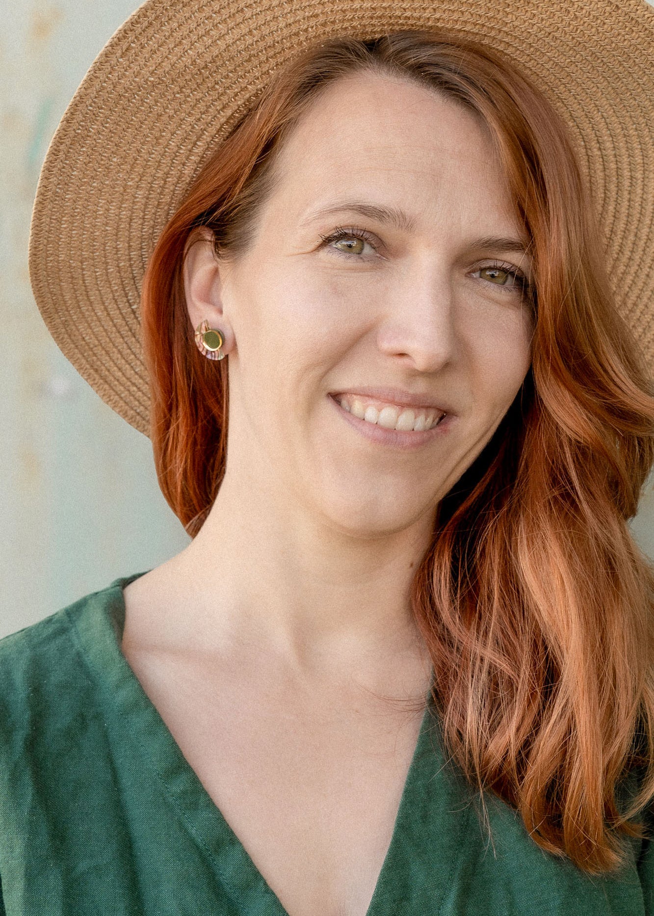 A woman with long auburn hair wears minimalist geometric stud earrings, a wide-brimmed straw hat, and a green dress, smiling softly while standing in front of a neutral-colored wall.