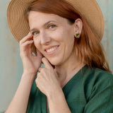A woman with reddish-brown hair wears a green dress, a straw hat, and minimalist geometric stud earrings. She smiles gently at the camera while touching her face, standing in front of a light-colored background.