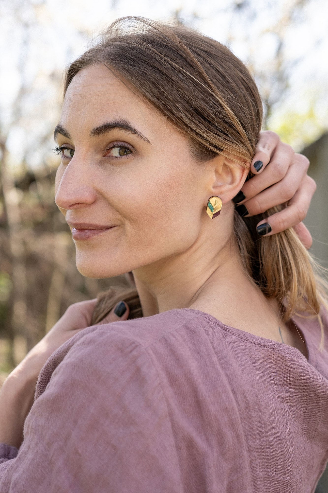 A woman with light brown hair in a low ponytail stands outdoors, wearing a mauve blouse and minimalist jewelry—gold leaf studs that catch the light. She looks over her shoulder at the camera, smiling softly, with one hand touching her hair.