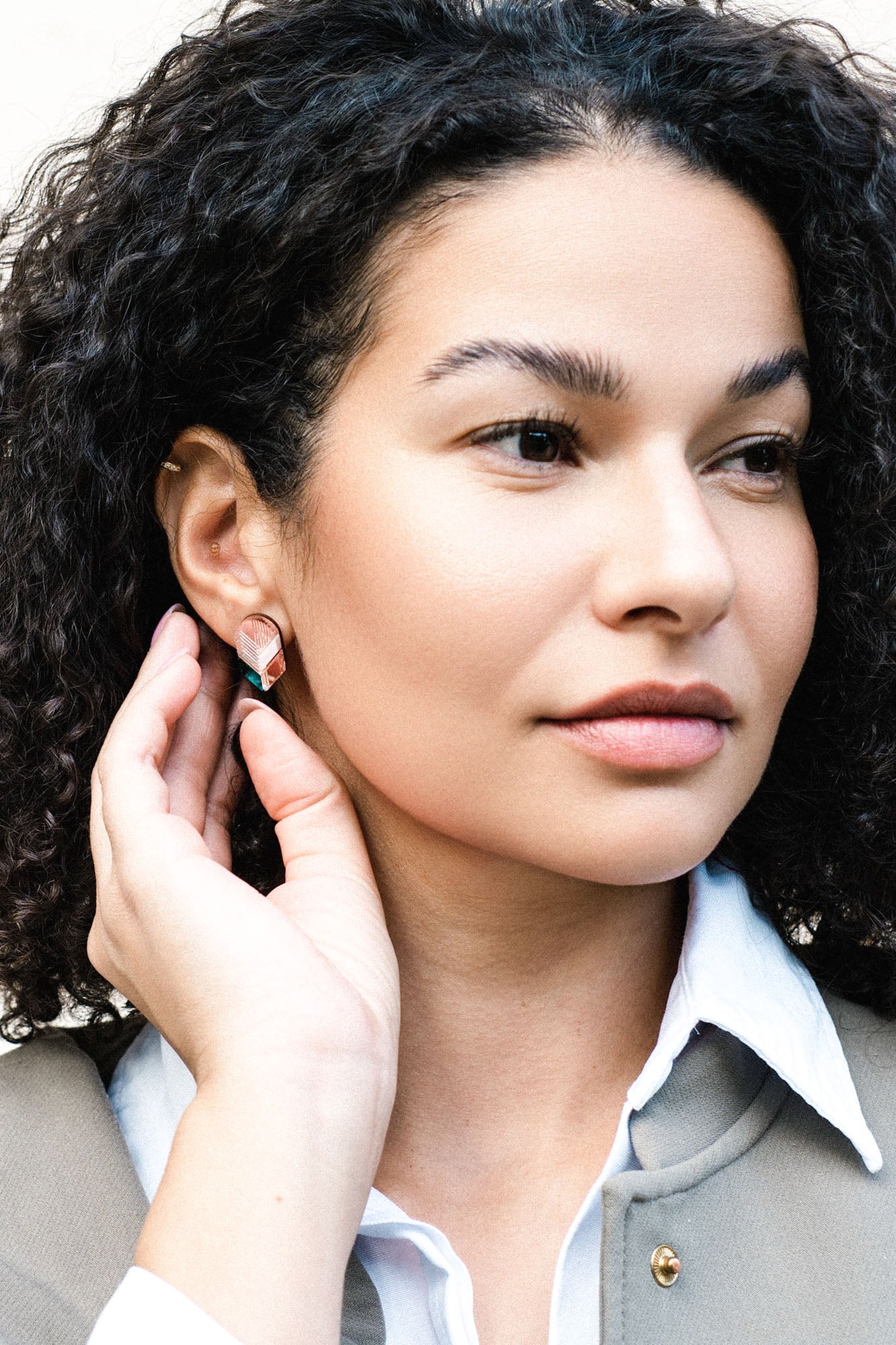 A woman with curly dark hair gazes to the side, gently touching her ear to show off handmade minimalist jewelry—delicate leaf studs. She wears a white collared shirt and a light gray jacket.