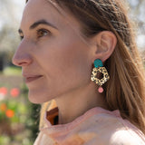 A woman with light brown hair gazes to the left, wearing a peach top and striking Leopard Earrings crafted from premium materials. The background is outdoors with blurred greenery and flowers.