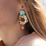 A close-up of a woman’s face in profile, wearing Leopard Earrings featuring timeless patterns—a large, colorful leopard print with a blue geometric shape and a small red drop—paired with a light peach top outdoors in sunlight.