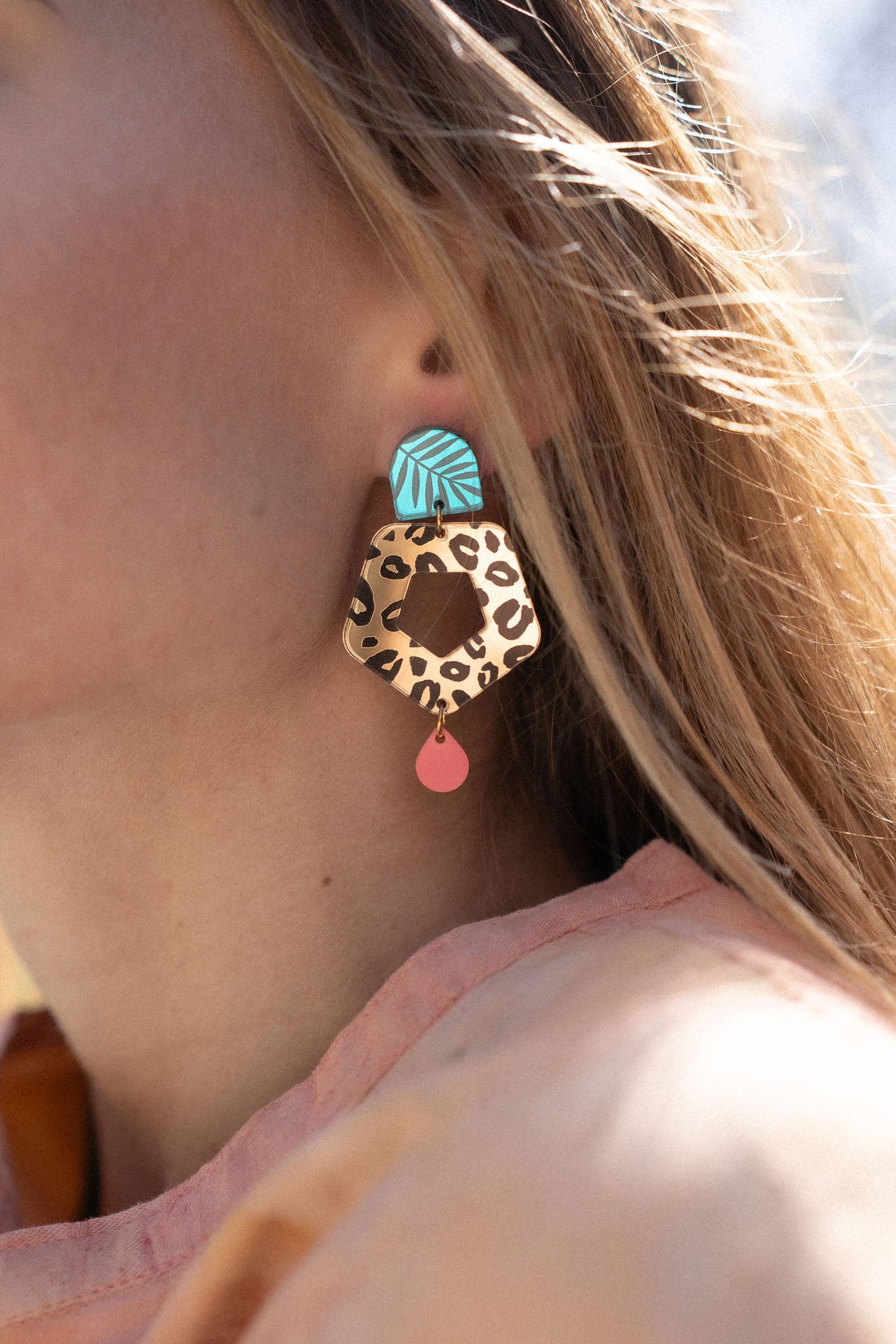 A close-up of a woman’s face in profile, wearing Leopard Earrings featuring timeless patterns—a large, colorful leopard print with a blue geometric shape and a small red drop—paired with a light peach top outdoors in sunlight.