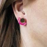 Close-up of a persons ear wearing lightweight Minimalist Geometric Stud Earrings, featuring a round gold stud and a pink, crescent-shaped accent. The person has straight brown hair tucked behind the ear.