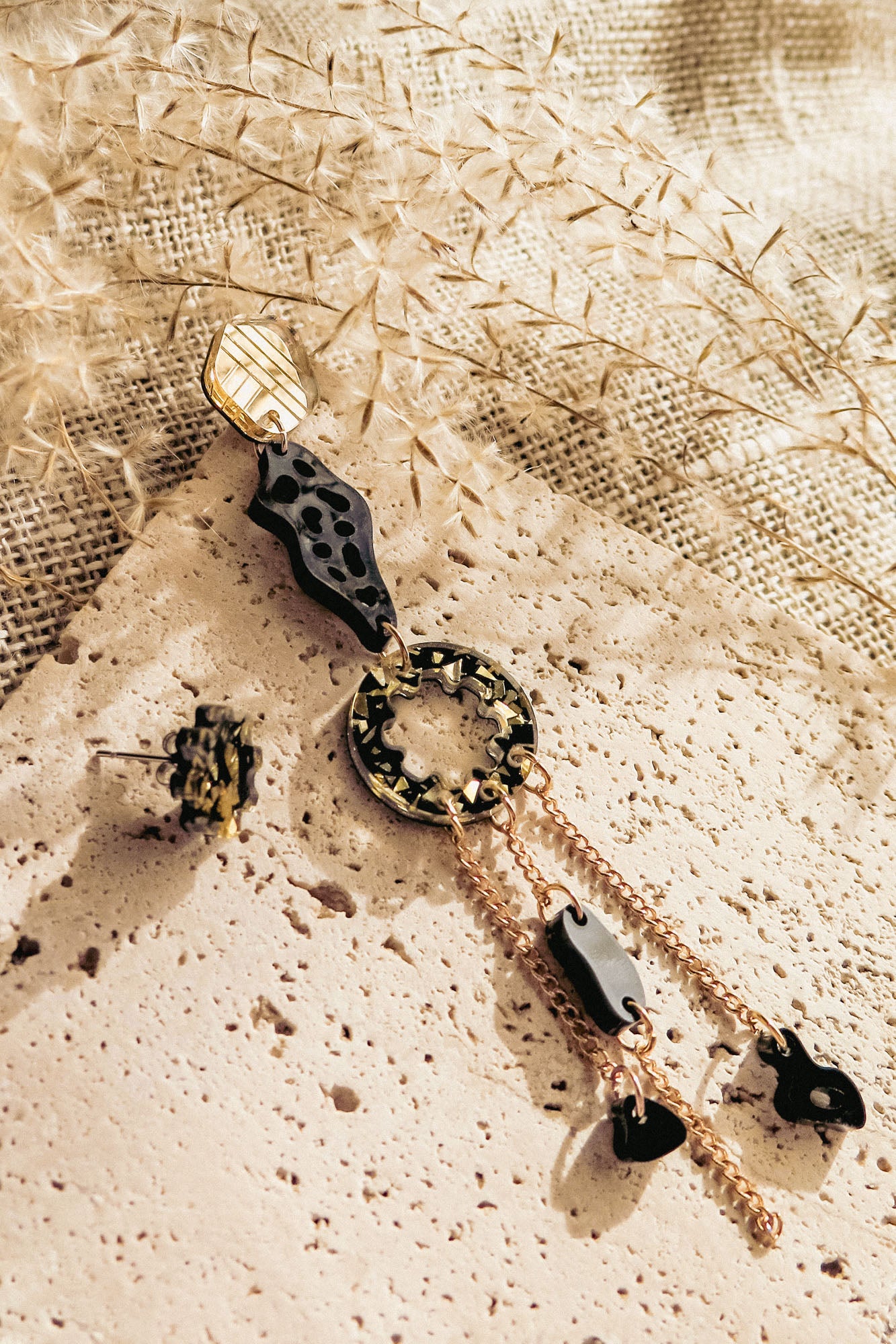 A modern, artistic mismatched extravagant earring in gold, black, and tortoiseshell rests on a textured stone surface, surrounded by dried foliage and beige woven fabric in soft natural light.