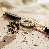 Close-up of a stylish gold bracelet and a matching floral-shaped ring with black and gold details, paired with lightweight handcrafted earrings, all arranged on a textured, light-colored stone surface in soft natural light.