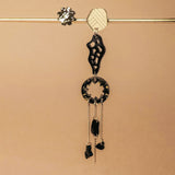 A single handcrafted earring featuring black and silver geometric shapes and dangling chains hangs from a horizontal metallic rod against a beige background—perfect for those who love lightweight earrings with an abstract flair.